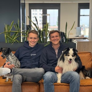 Lasse Steffen und Marco Kippler sitzen als Gründer mit blauen Hoodies auf einer Couch, schauen lachend in die Kamera und haben beiden jeweils einen Hund im Arm.