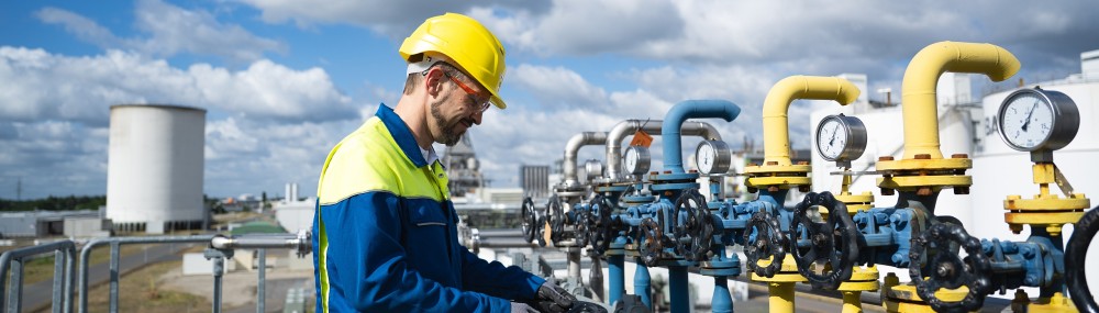 Ein Mann mit Sicherheitshelm im Arbeitrsanzug steht im Freien an einer Großindustrieanlage mit Rohren. Im Hinterrgrund ein Kühlturm.