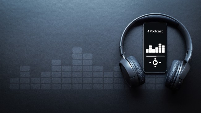 Schwarze Kopfhörer, dazwischen ein Smarthphone. Auf dem Display steht: Podcast. Alles vor schwarzem Hintergrund. Grafisch inzeniertes Bild.
