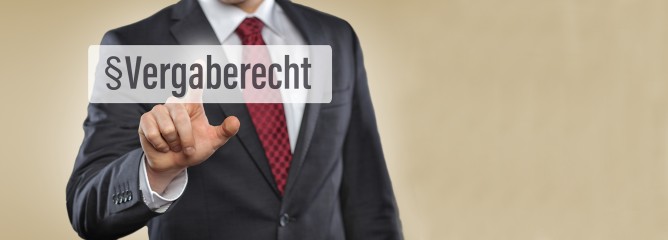 Mensch im Anzug tippt mit dem Finger auf das im Vordergrund dargestellte Wort 'Vergaberecht' 