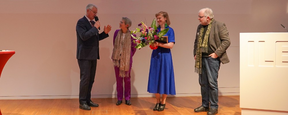 Es handelt sich um die Verleihung der Auszeichnung des ILB-Kunstpreis 2022. Es sind vier Personen. Eine hält einen Blumenstrauß und eine Urkunde, eine andere hält ein Mikrofon, während sie spricht. Zwei andere Personen hören aufmerksam zu.