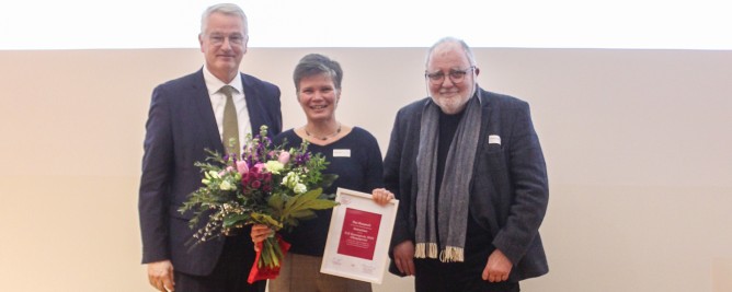 Es handelt sich um die Verleihung einer Auszeichnung. Es sind vier Personen. Eine hält einen Blumenstrauß, eine andere hält ein Mikrofon, während sie spricht. Eine andere Person hört aufmerksam zu und eine weitere Person zeigt eine Urkunde. Im Hintergrund sieht man das Porträt einer Künstlerin und das Logo des Preises, auf dem "ILB-Kunstpreis Brandenburg 2022" steht.
