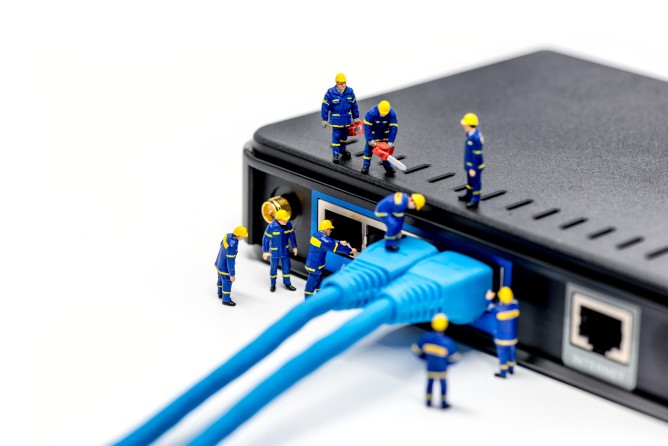 Ein Team Techniker bestehend aus vielen Mini-Figuren schließen ein Netzwerkkabel an einen Router an.